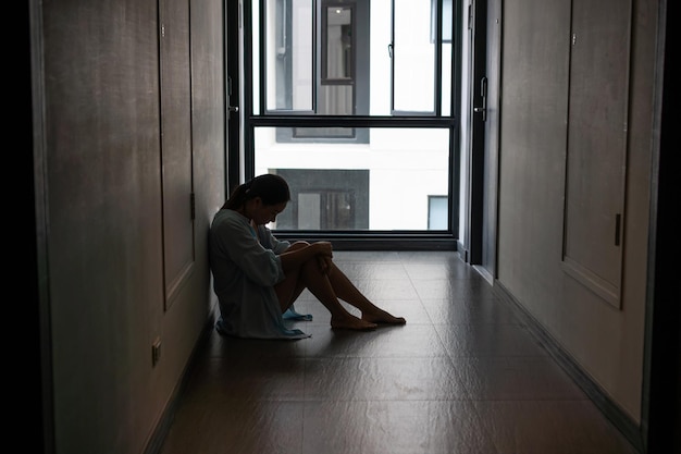 Stressée jeune femme asiatique souffrant de dépression et assise seule à l'extérieur de la chambre ou du couloir de l'appartement Triste malheureuse déçue de la violence domestique ou arrêter la violence et le concept de viol Copier l'espace