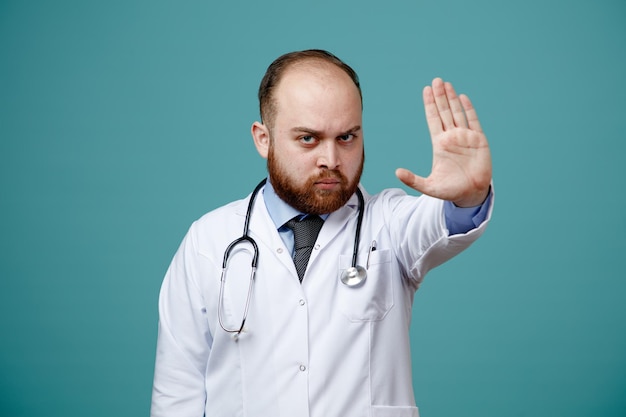 Strict jeune médecin de sexe masculin portant un manteau médical et un stéthoscope autour du cou regardant la caméra montrant un geste d'arrêt isolé sur fond bleu