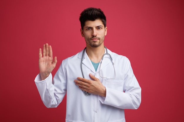 Strict jeune médecin de sexe masculin portant un uniforme médical et un stéthoscope autour du cou regardant la caméra en gardant la main sur la poitrine montrant le geste d'arrêt isolé sur fond rouge