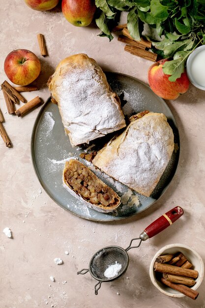 Strudel aux pommes classique en tranches fait maison avec du sucre glace sur une plaque en céramique avec des pommes fraîches, des feuilles vertes et des bâtons de cannelle au-dessus.