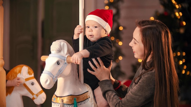 Studio décoré de Noël une jeune mère souriante debout près de son bébé sur le carrousel