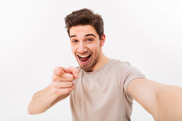 Studio photo d'homme positif en t-shirt décontracté et poils sur le visage souriant et pointant le doigt sur la caméra tout en prenant selfie, isolé sur mur blanc