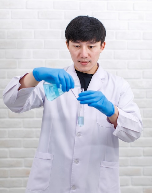 Studio de portrait tourné Scientifique masculin professionnel asiatique en blouse de laboratoire blanche gants en caoutchouc debout souriant regarder la caméra tenant la verrerie de fiole Erlenmeyer vide vide dans les mains sur fond de mur de briques.