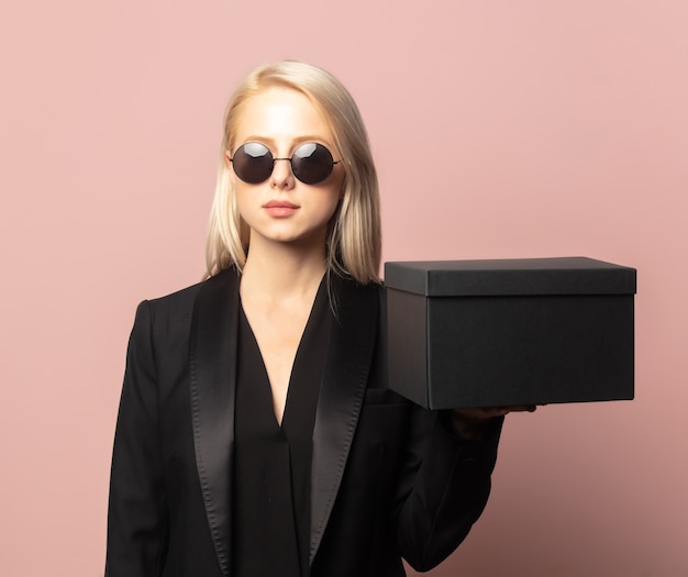 Style blonde en blazer et lunettes de soleil avec boîte-cadeau noire sur rose