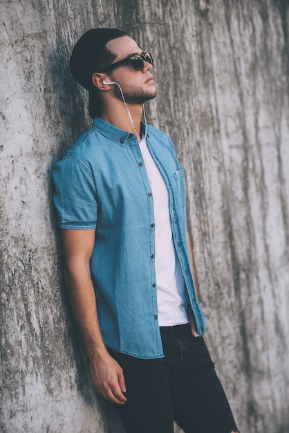 Style décontracté. Beau jeune homme en casquette et lunettes de soleil écoutant de la musique tout en portant des écouteurs et en se penchant sur le mur de béton