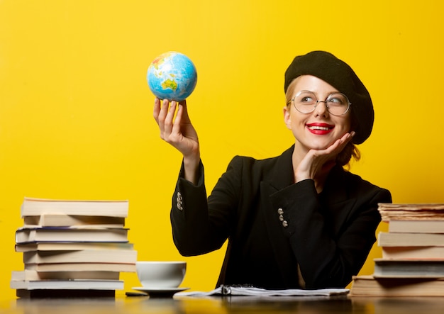 Style femme blonde en béret assis à table avec des livres autour et détient globe sur jaune