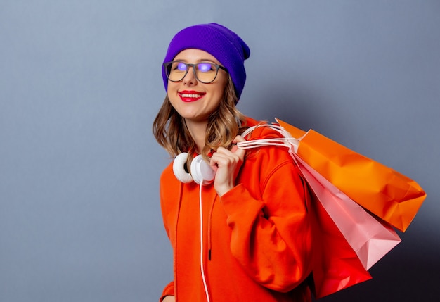 Style fille en capuche orange et chapeau violet avec des sacs à provisions sur mur gris