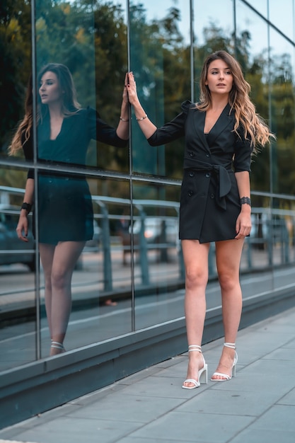 Style de rue, une jeune femme caucasienne blonde entreprenante entrant dans le bureau pour travailler dans un costume noir et des talons. Reflété dans le verre du bâtiment en verre