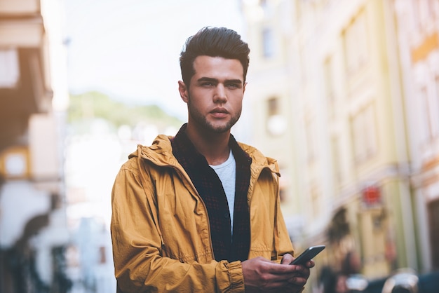 Style de ville. Beau jeune homme tenant un téléphone intelligent et regardant loin en se tenant debout dans la rue de la ville