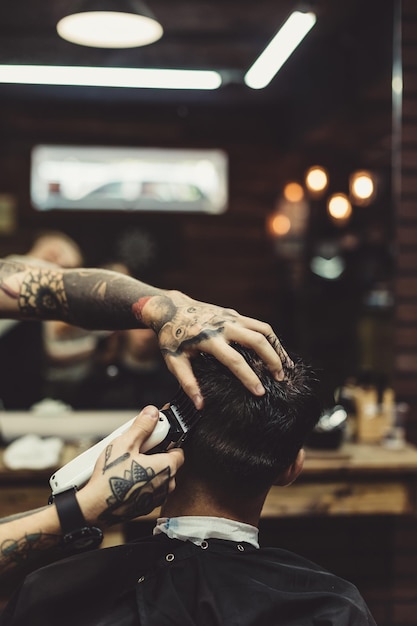 Styliste tatoué concentré sur le rasage de l'homme avec une machine à faire la coiffure