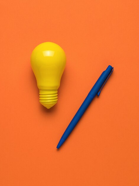 Un stylo bleu et une ampoule jaune vif sur fond orange. Mise à plat.