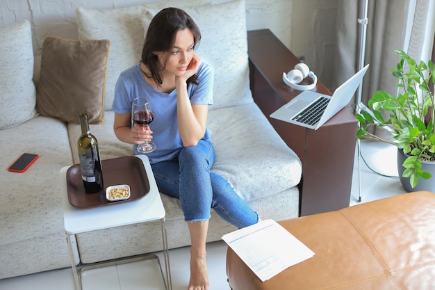 Succès belle jeune femme assise sur un canapé dans le salon en buvant du vin rouge