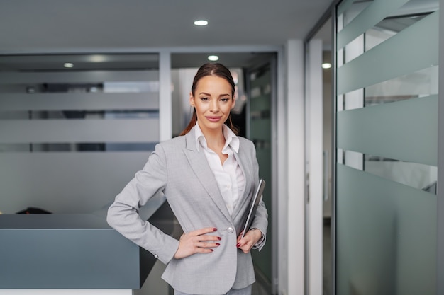 Succès caucasien jeune femme d'affaires debout à l'intérieur de l'entreprise, tenant la main sur la hanche et dans l'autre main tenant la tablette.