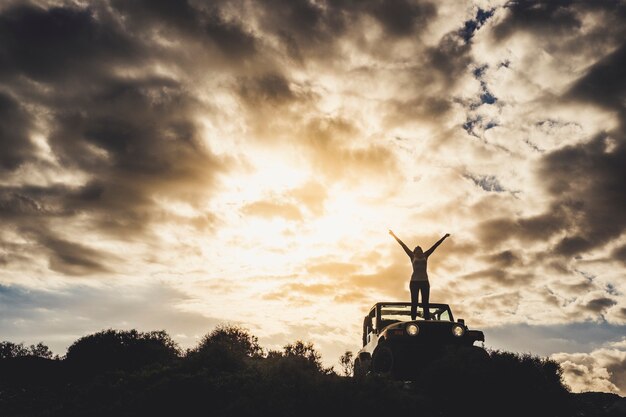 Succès et concept de voyageur solitaire avec une fille en silhouette debout sur sa voiture et ouvrant les bras jusqu'au ciel dramatique d'un coucher de soleil doré