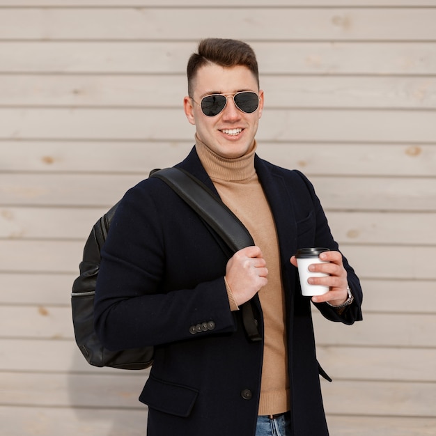 Succès jeune homme avec un beau sourire dans des lunettes élégantes dans un manteau élégant dans un pull tricoté en jeans à la mode tient dans sa main une tasse de délicieux café