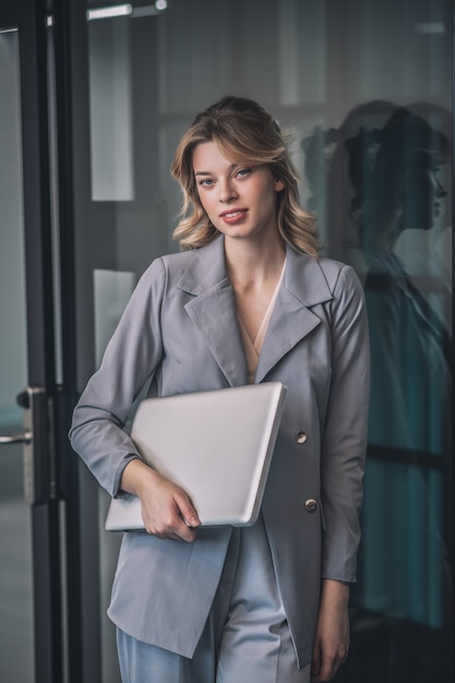 Succès, perspective. Femme d'affaires jeune blonde réussie en costume gris tenant un ordinateur portable dans la main debout près de la porte en verre au bureau