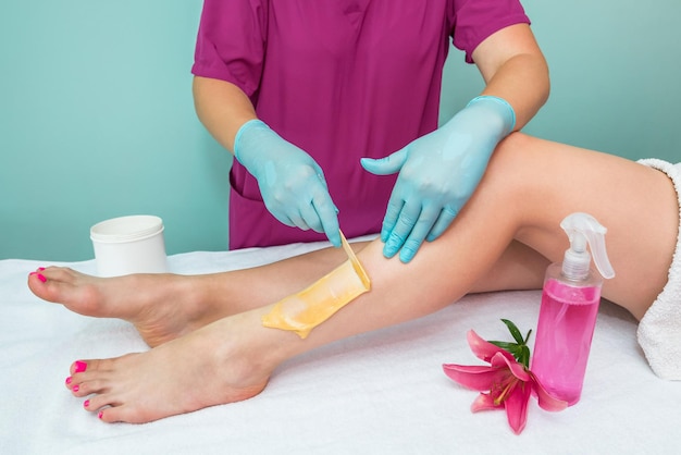 Sucrage des jambes Une esthéticienne fait une épilation en pâte à sucre des jambes d'une femme dans un salon de beauté Cosmétologie esthétique féminine Appliquer la pâte à sucre avec une spatule en bois