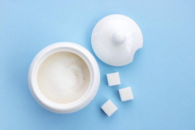 Sucrier ouvert avec du sucre à côté de cubes de sucre raffiné sur fond bleu