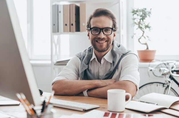 Super journée de travail ! Beau jeune homme regardant la caméra et souriant alors qu'il était assis sur un lieu de travail dans un bureau créatif