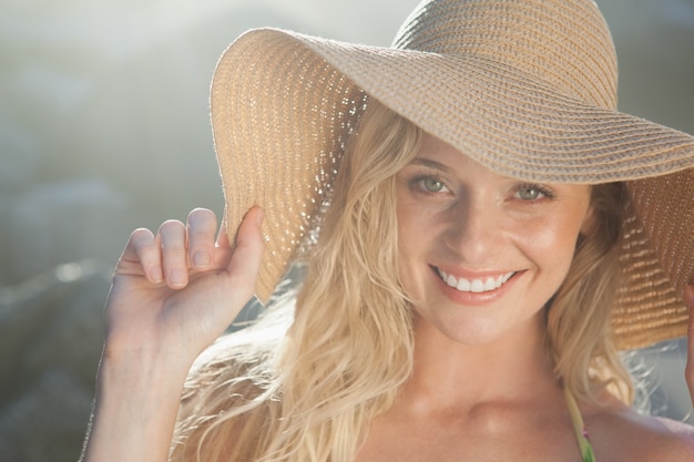 Superbe blonde au chapeau de paille souriant à la caméra sur la plage