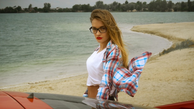 Superbe blonde à lunettes se tient à la voiture rouge au bord de la mer
