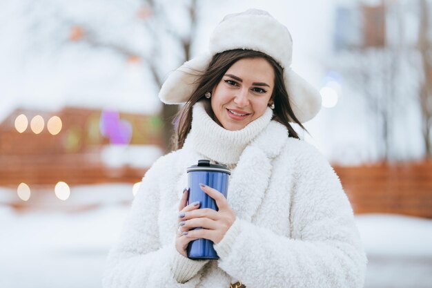 Superbe brune caucasienne jeune femme en manteau de fourrure blanche et chapeau tient un thermos avec du thé