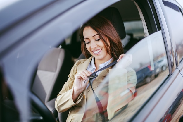 Superbe brunette caucasienne habillée décontractée chic assise dans sa voiture et attacher la ceinture de sécurité.