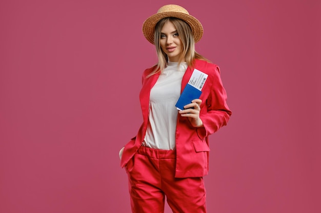 Superbe dame blonde en chapeau de paille, chemisier blanc et tailleur-pantalon rouge. Elle a mis sa main dans la poche, souriante, tenant un passeport et un billet, posant sur fond rose. Concept de voyage. Gros plan, espace de copie