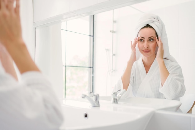 Superbe femme adulte confiante portant une serviette sur la tête assise et vérifiant sa mâchoire devant le miroir dans la salle de bain à la maison Une jolie femme séduisante mode de vie beauté et concept cosmétique