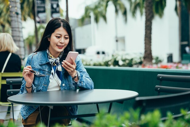 superbe femme asiatique qui enlève des lunettes de soleil pour lire un message téléphonique dans un coin salon extérieur. une dame souriante en smart casual parcourt les flux de médias sociaux pendant la pause déjeuner. style de vie authentique