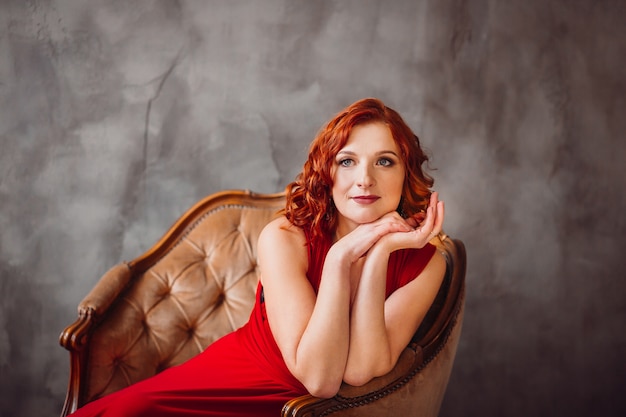 Superbe femme aux cheveux rouges en longue robe rouge se trouve dans la douce chaise devant la fenêtre