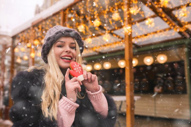 Une superbe femme blonde porte un chapeau tricoté tenant un biscuit de pain d'épice sucré à la foire de Noël. Espace pour le texte