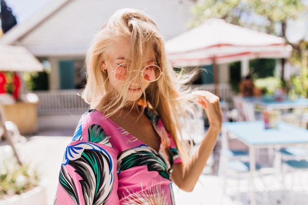 Superbe femme bronzée dans des verres roses passant du temps à la station balnéaire Photo en plein air d'un incroyable modèle féminin aux cheveux longs en tenue lumineuse profitant d'une journée ensoleillée