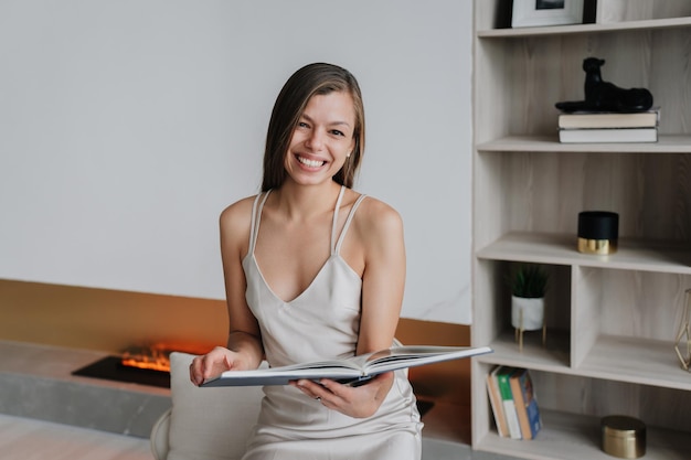 Superbe femme brune gaie en robe de soirée tient un livre ouvert regarde la caméra est assis sur une chaise contre une étagère et une cheminée à la maison Une femme caucasienne réussie lisant à la maison