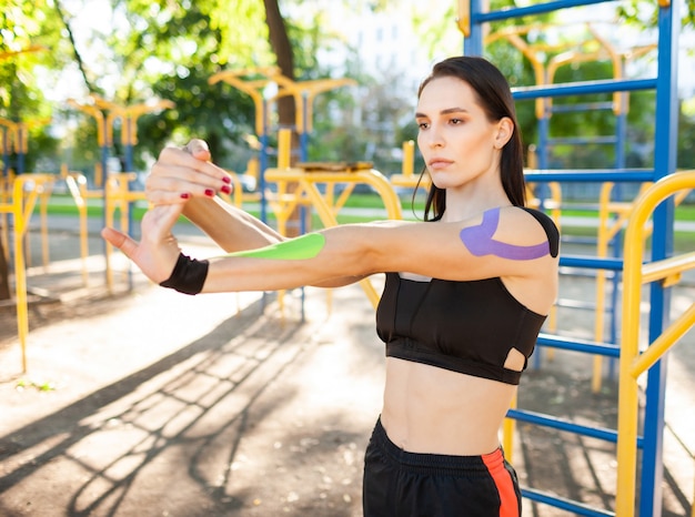 Superbe femme brune musclée flexible portant une tenue de sport noire, bras qui s'étend. Jeune athlète féminine confiante pratiquant la gymnastique, l'échauffement, le kinesiotaping coloré sur le corps.