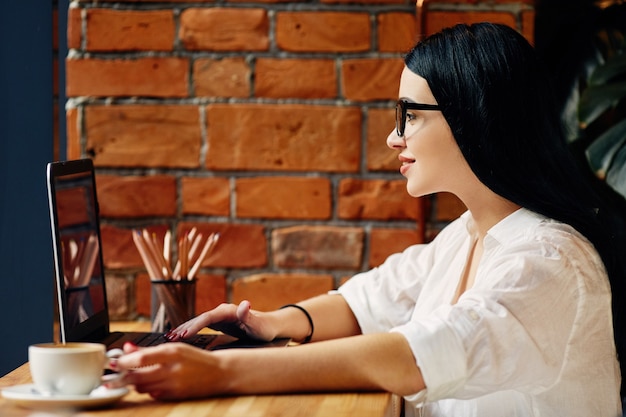 Superbe fille aux cheveux noirs portant des lunettes assis dans un café avec ordinateur portable et tasse de café, concept indépendant, portrait, vêtu d'une chemise blanche.