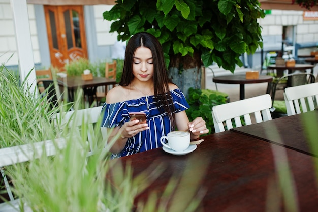 Superbe fille brune assise sur la table au café avec une tasse de café et utilisant le téléphone