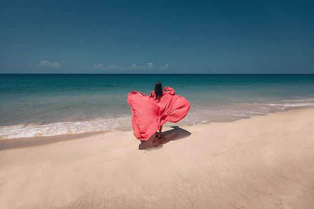 Superbe fille sur la plage exotique ensoleillée au bord de l'océan. La jeune femme porte une robe rouge étonnante flottant dans la brise légère, vue arrière ; notion de mode.