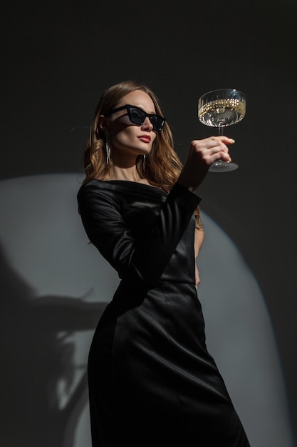 Superbe jeune femme chic à la mode avec des lunettes de soleil cool dans une élégante robe noire élégante avec une coupe de champagne vintage sur fond sombre lors d'une fête Jolie dame célèbre un événement