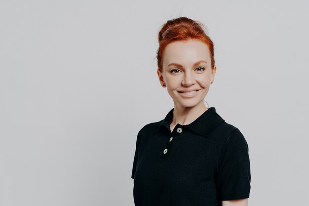 Superbe jeune femme confiante des années 30 aux cheveux rouges en chignon posant isolée sur fond gris studio, vêtue avec désinvolture d'un t-shirt noir, regardant joyeusement la caméra et exprimant sa confiance