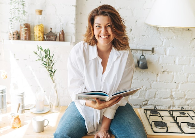 Superbe jeune femme heureuse plus corps de taille positif en chemise blanche lisant un livre de cuisine dans la cuisine