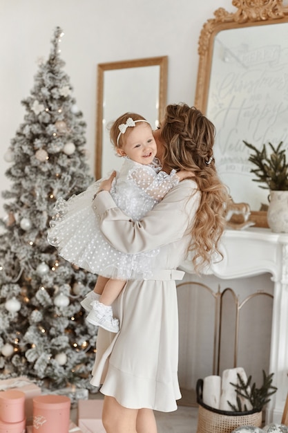 Superbe jeune femme en robe élégante posant avec sa petite fille en robe moelleuse dans un intérieur décoré de Noël.