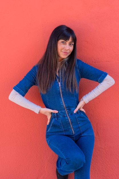 Superbe jeune fille brune souriante appuyée contre un mur dans une tenue en denim bleu. Jolie caucasienne posant sur fond rouge