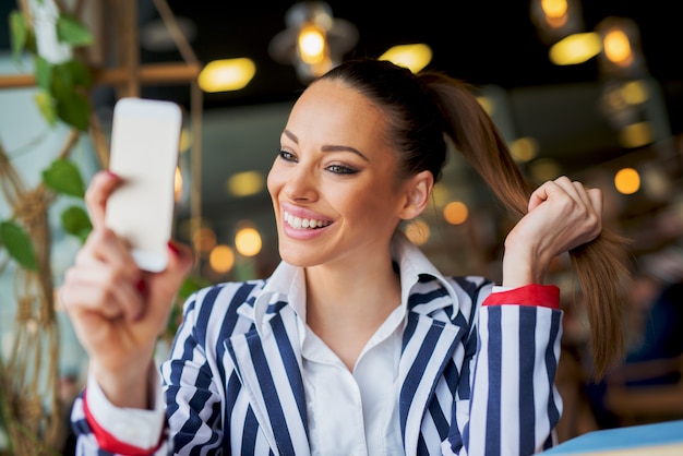 Superbe jeune fille élégante fait un selfie dans un café tout en tenant sa queue de cheval.