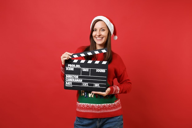 Superbe joyeuse jeune fille de Santa en chapeau de Noël tenant un film noir classique faisant un clap isolé sur fond rouge. Bonne année 2019 concept de fête de vacances célébration. Maquette de l'espace de copie.
