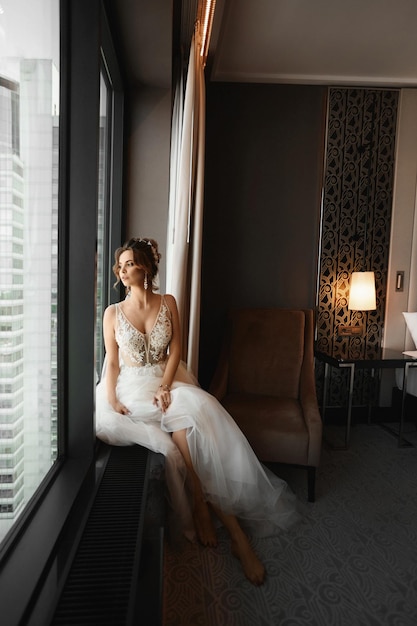 Superbe mariée dans une robe de mariée à la mode et des boucles d'oreilles luxueuses assises à côté de la fenêtre