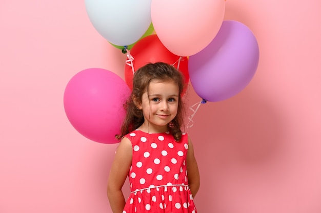 Superbe petite fille vêtue d'une robe d'été à pois, tenant des ballons multicolores derrière son dos, regardant la caméra. Concept de la journée de la femme et de la fille heureuse. Anniversaire, festivals événements, célébrations.