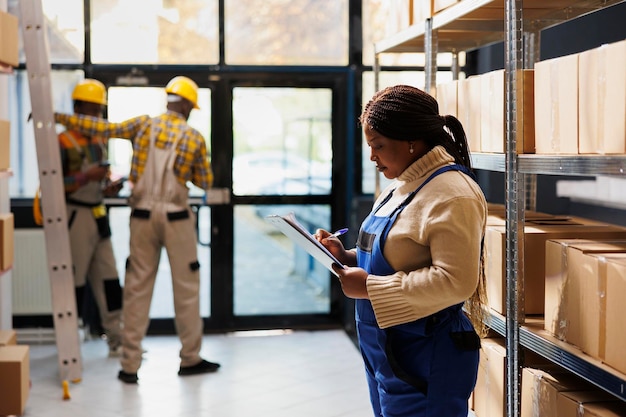 Superviseur d'approvisionnement de stock d'entrepôt afro-américain écrivant sur le presse-papiers. Opérateur d'entrepôt de vente au détail femme debout près de l'étagère des boîtes en carton et prenant des notes sur la gestion des stocks