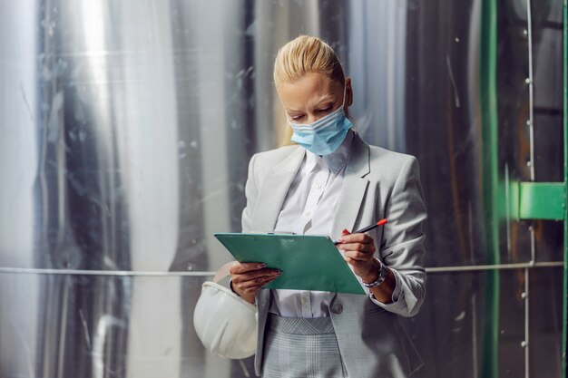 Superviseur blonde dévouée et réussie en tenue de soirée avec masque facial de protection remplissant la documentation tout en se tenant dans une centrale électrique pendant un coronavirus.