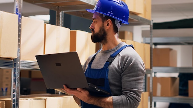 Superviseur de dépôt tenant un ordinateur portable avec la liste logistique des stocks, regardant les packs de marchandises et planifiant la distribution des commandes. Employé masculin utilisant un ordinateur pour effectuer un contrôle de qualité. Prise de vue à main levée.